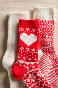 Three knit socks arranged on a beige fabric background: one white sock, one red-and-white heart pattern sock, and one red-and-white Nordic pattern sock.