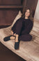 A woman sits on a beige upholstered daybed wearing a dark plum pajama set and dark slip-on slippers with brown soles in a cozy indoor setting.
