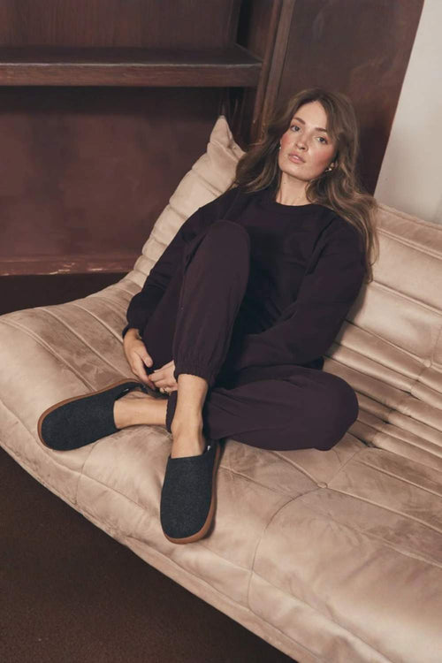 A woman sits on a beige upholstered daybed wearing a dark plum pajama set and dark slip-on slippers with brown soles in a cozy indoor setting.