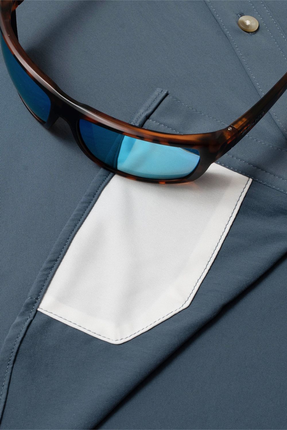 Close-up of a slate blue button-up shirt with a white chest pocket and tortoiseshell sunglasses resting on top.