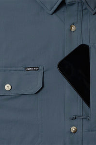 Close-up of a slate blue button-up shirt pocket with a visible PONCHO label, a black smartphone tucked into the chest pocket, and metallic buttons.