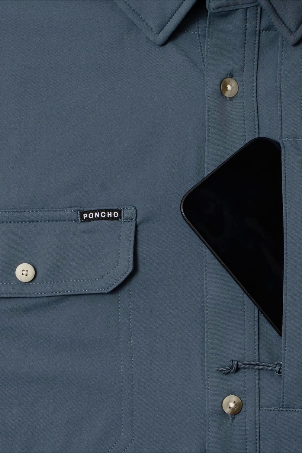 Close-up of a slate blue button-up shirt pocket with a visible PONCHO label, a black smartphone tucked into the chest pocket, and metallic buttons.