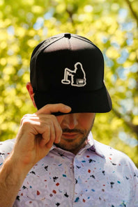 A man outdoors wearing a black baseball cap with a white embroidered graphic.  He is wearing a white button-down shirt with a multicolor confetti pattern, set against a bright, sunlit background of green bokeh trees.