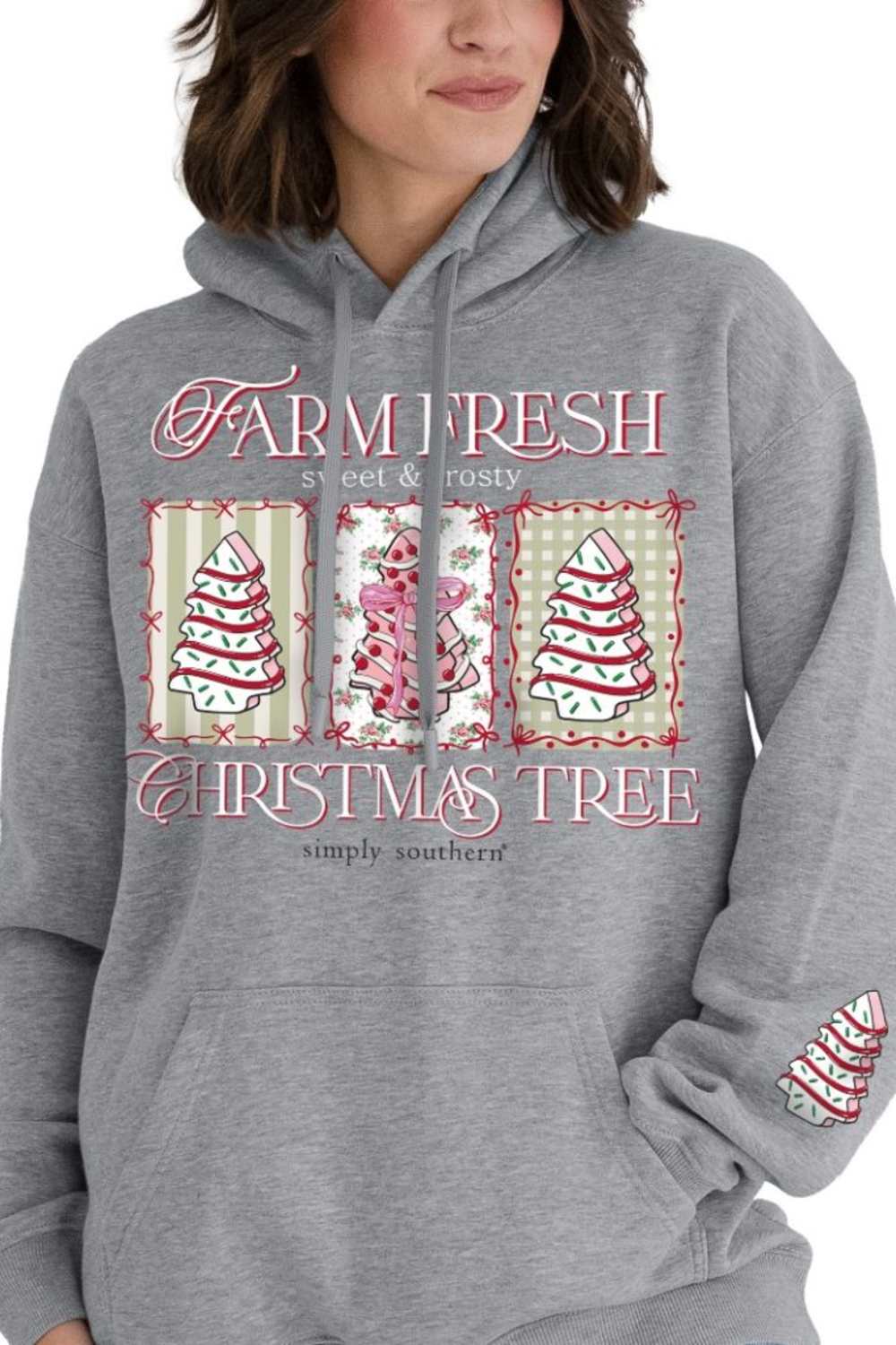 A woman wearing a heather gray pullover hoodie with a festive Christmas graphic on the front. The graphic features the words 'FARM TO FRESH' and 'CHRISTMAS TREE' in pink script, with three vertical panels showing decorated Christmas trees and a pink ornament.