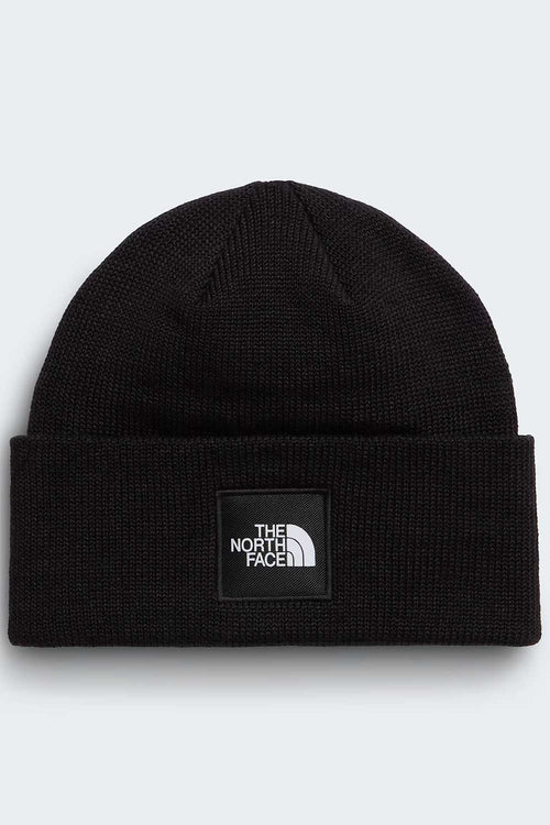 A black ribbed knit beanie hat featuring a black square patch on the front with the white 'THE NORTH FACE' logo, set against a light gray background.