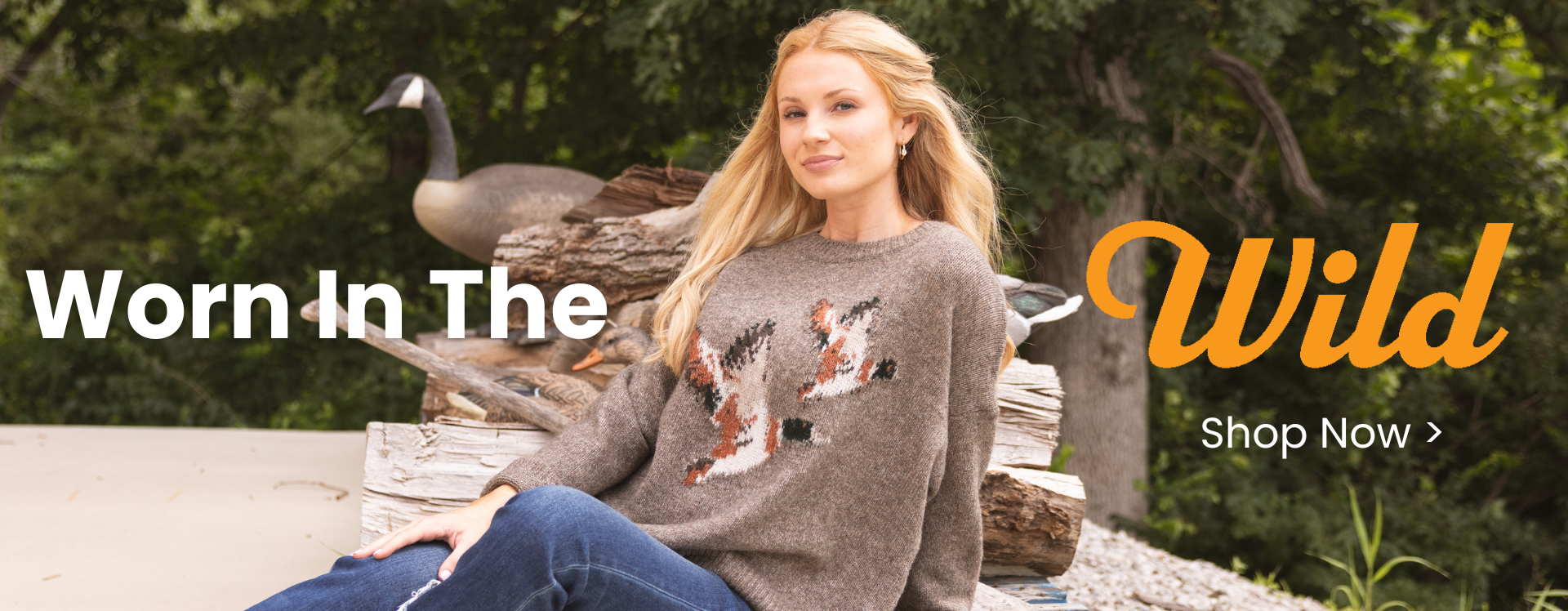 Promotional banner featuring a model seated outdoors wearing a brown sweater with a wildlife-inspired pattern and blue jeans. Large text reads, "Worn In The Wild," alongside a "Shop Now" call-to-action, with rustic logs and duck decoys in the background.