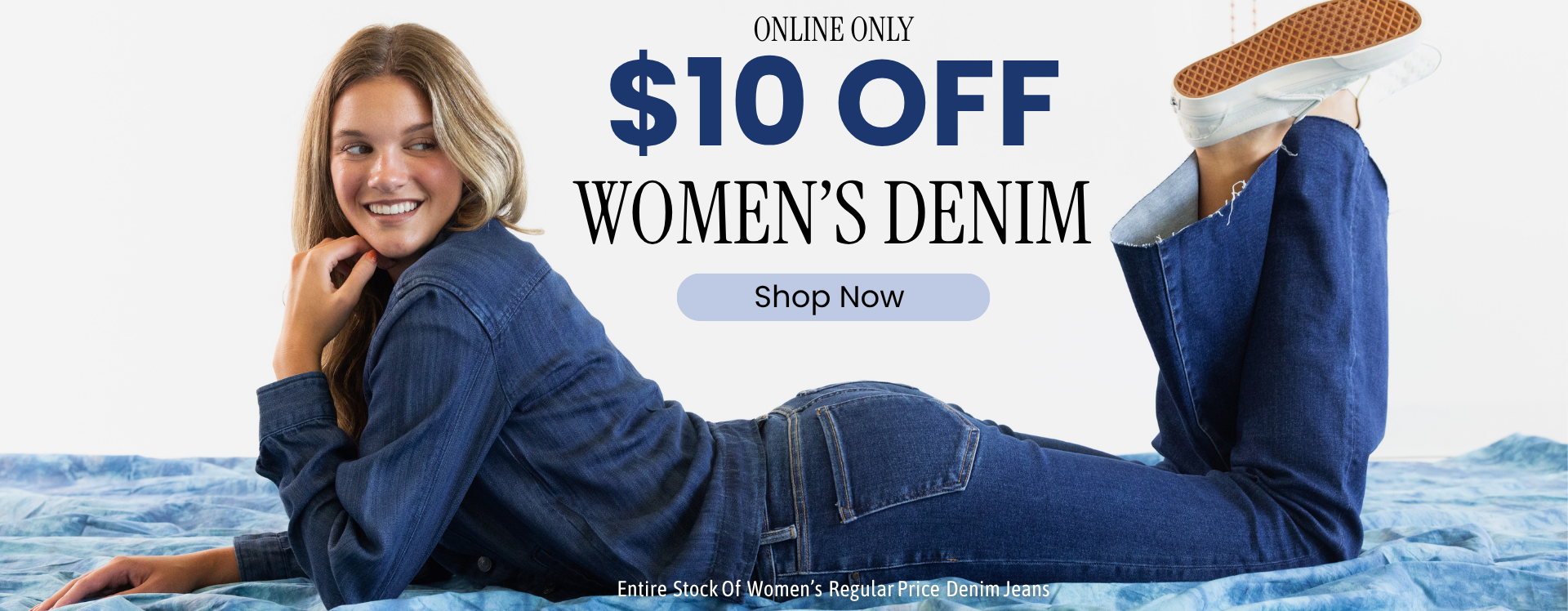 Promotional banner of a women laying on a tie-dye blue sheet with a clean white background on her stomach with her knees bent and feet kicked up behind her. She wearing dark wash flare denim jeans and a dark wash denim shirt and white sneakers. Text reads ONLINE ONLY $10 OFF WOMEN'S DENIM. A light blue call-to-action reads SHOP NOW. Fine print text reads ENTIRE STOCK OF WOMEN'S REGULAR PRICE DENIM JEANS