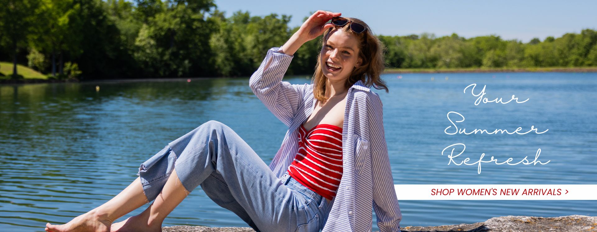 Girl sitting in front of a lake on top of a rock surface wearing a red and white striped strapless top, a white and blue stripe shirt, & jeans and text that reads "your summer refresh - shop women's new arrivals"