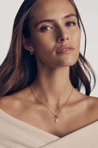 A close-up portrait of a woman wearing a delicate gold necklace with a small teardrop-shaped clear gemstone pendant, styled with a cream off-the-shoulder top against a light gray background.
