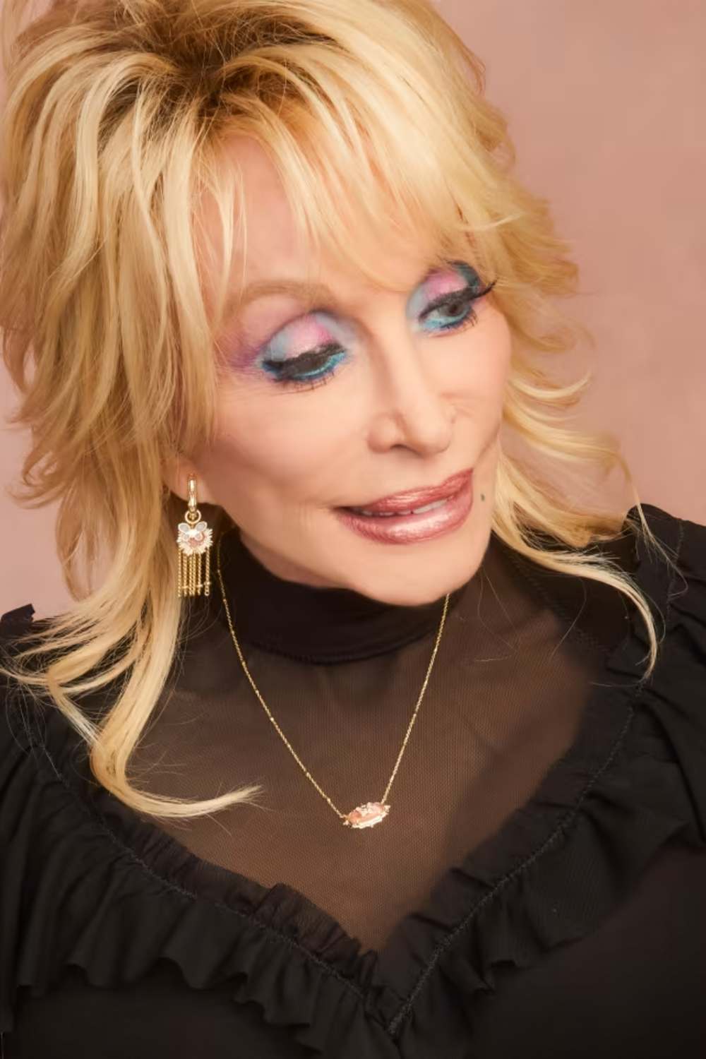 A close-up portrait of a blonde woman wearing a black ruffled turtleneck top, gold jewelry including a necklace and dangling earrings, and colorful blue and purple eyeshadow, set against a solid dusty pink background.