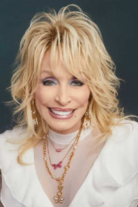 A close-up portrait of a smiling blonde woman with voluminous hair, wearing layered gold necklaces, dangling earrings, and a white top against a dark blue-gray background.