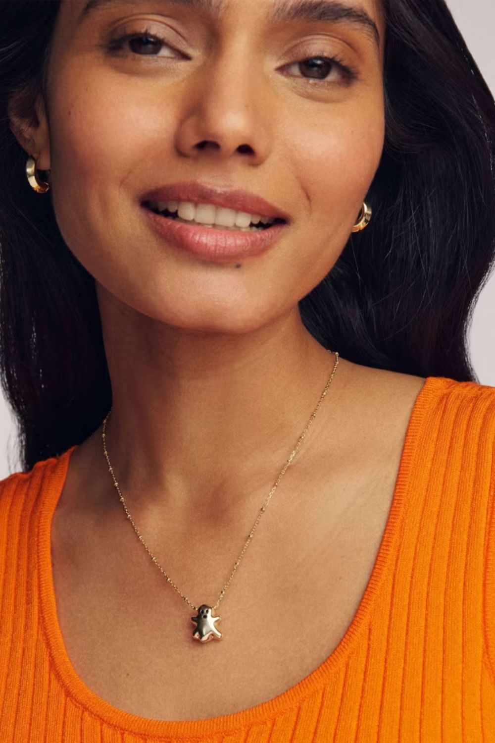 A close-up of a woman wearing a delicate gold chain necklace with a small gold ghost pendant, paired with a bright orange ribbed top.