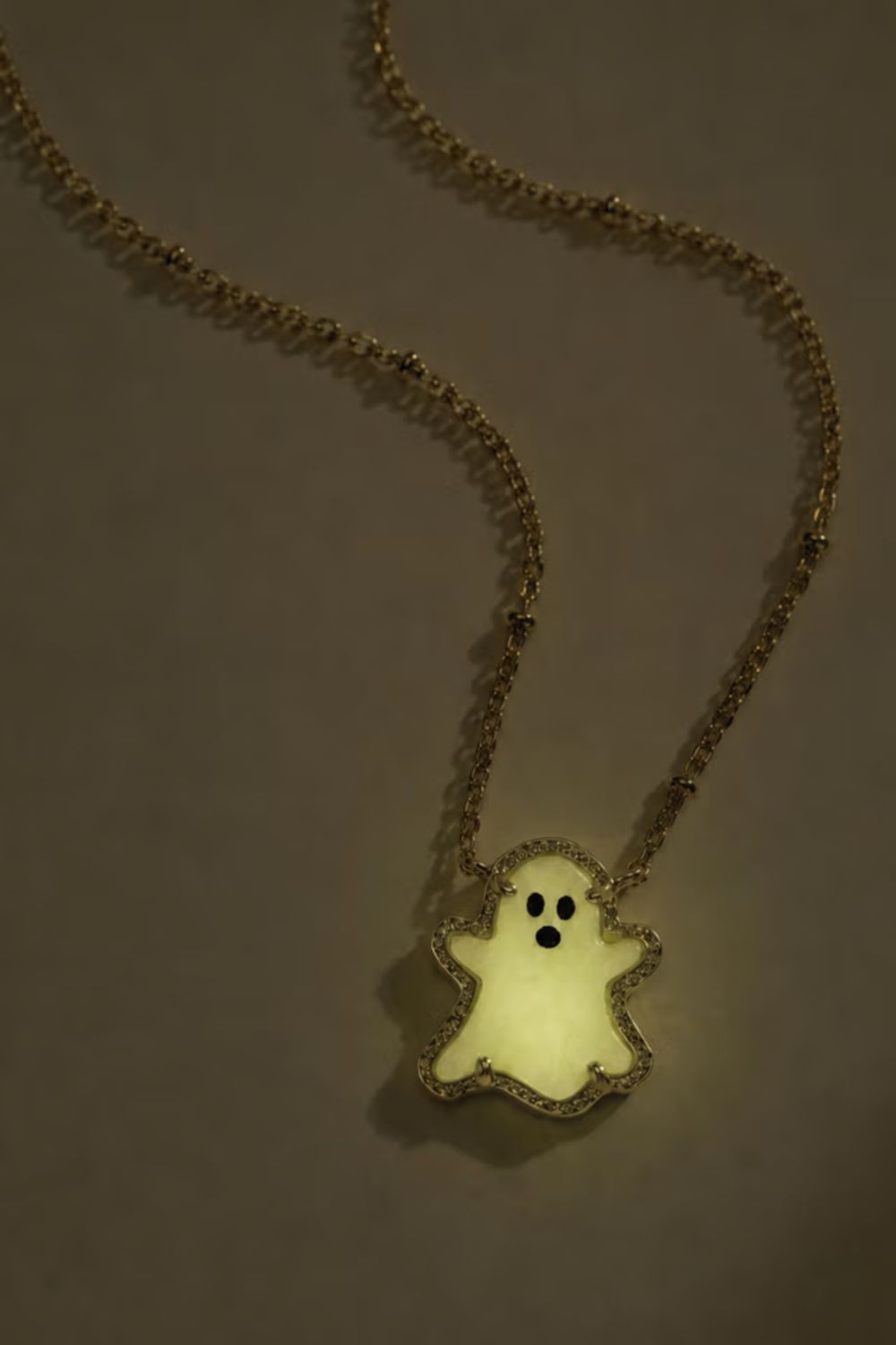 A gold chain necklace with a glowing, ghost-shaped pendant, photographed against a dark gray background.