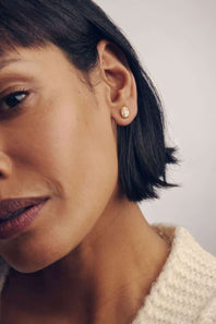 A close-up of a woman wearing a small gold stud earring with a clear gemstone, shown against a soft light gray background.