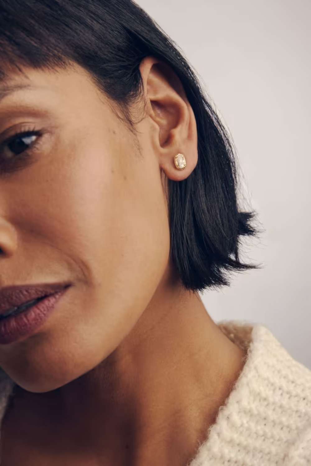 A close-up of a woman wearing a small gold stud earring with a clear gemstone, shown against a soft light gray background.
