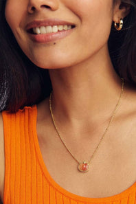A close-up lifestyle shot of a woman wearing a delicate gold chain necklace with a small pumpkin oval pendant, paired with a bright orange ribbed tank top.
