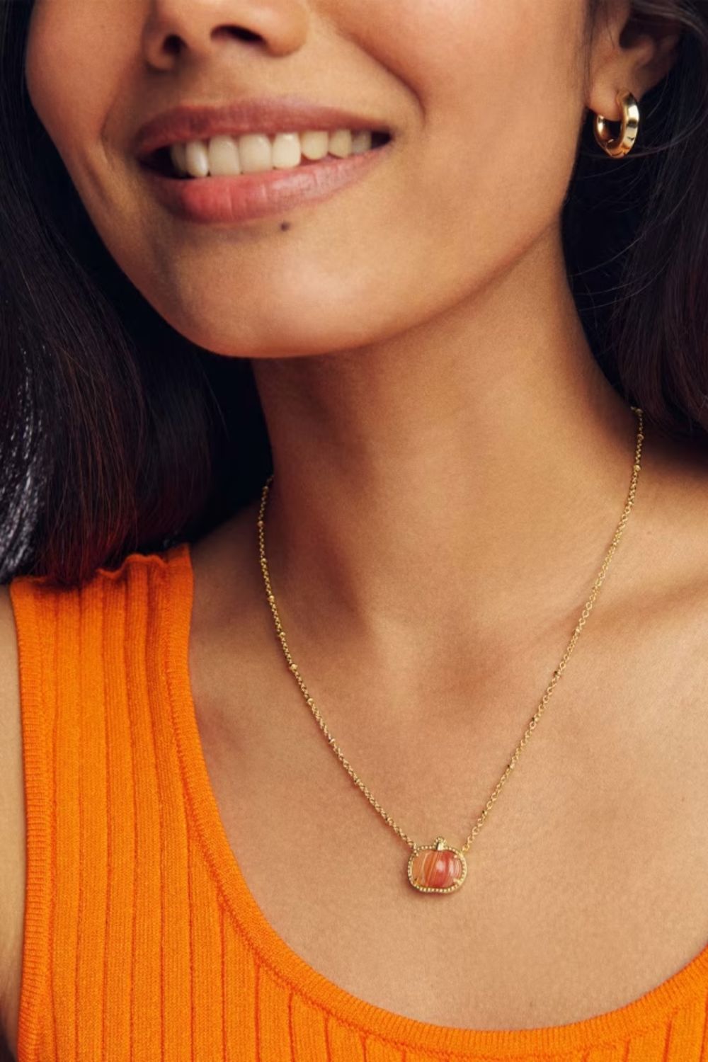 A close-up lifestyle shot of a woman wearing a delicate gold chain necklace with a small pumpkin oval pendant, paired with a bright orange ribbed tank top.