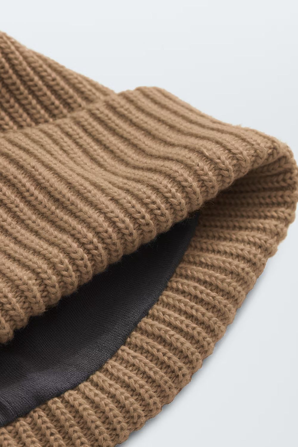 Close-up of a ribbed knit beanie in a warm camel brown color, showing the texture and a black inner lining on a light gray background.