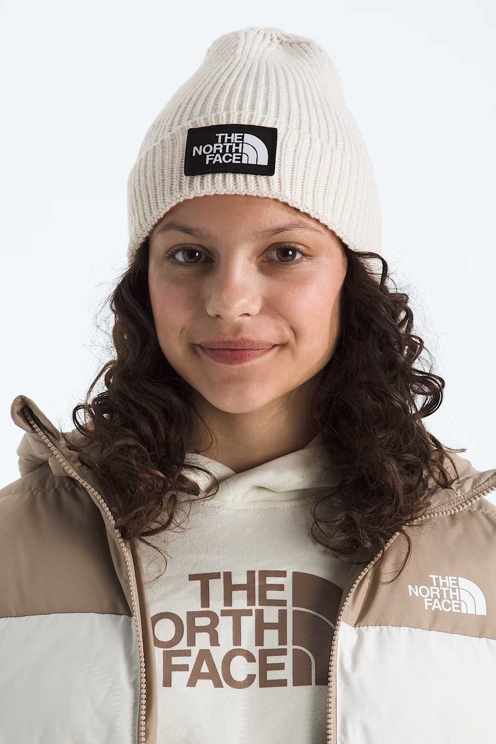 A young girls wearing an off-white ribbed knit beanie hat featuring a black rectangular patch on the front with the white 'THE NORTH FACE' logo.