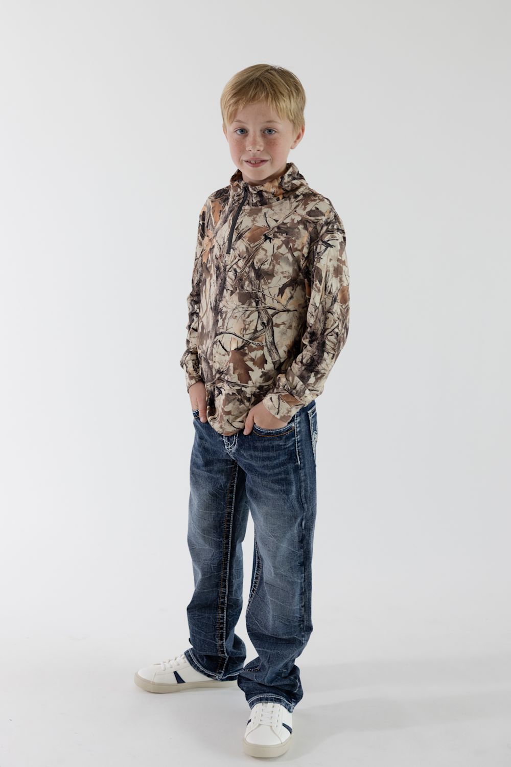 A full-length front view of a young boy with short blonde hair smiling at the camera, wearing a long-sleeve quarter-zip pullover in Leaf Camo with his hands tucked into the pockets of dark wash blue jeans, completed with white sneakers on a solid white background.