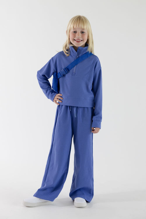 A full front view of a young girl with blonde hair smiling and standing hand-on-hip, wearing a bluish-purple ribbed quarter-zip pullover, matching wide-leg pants, and white sneakers, styled with a matching Perla Blue belt bag slung across her chest against a plain light background.