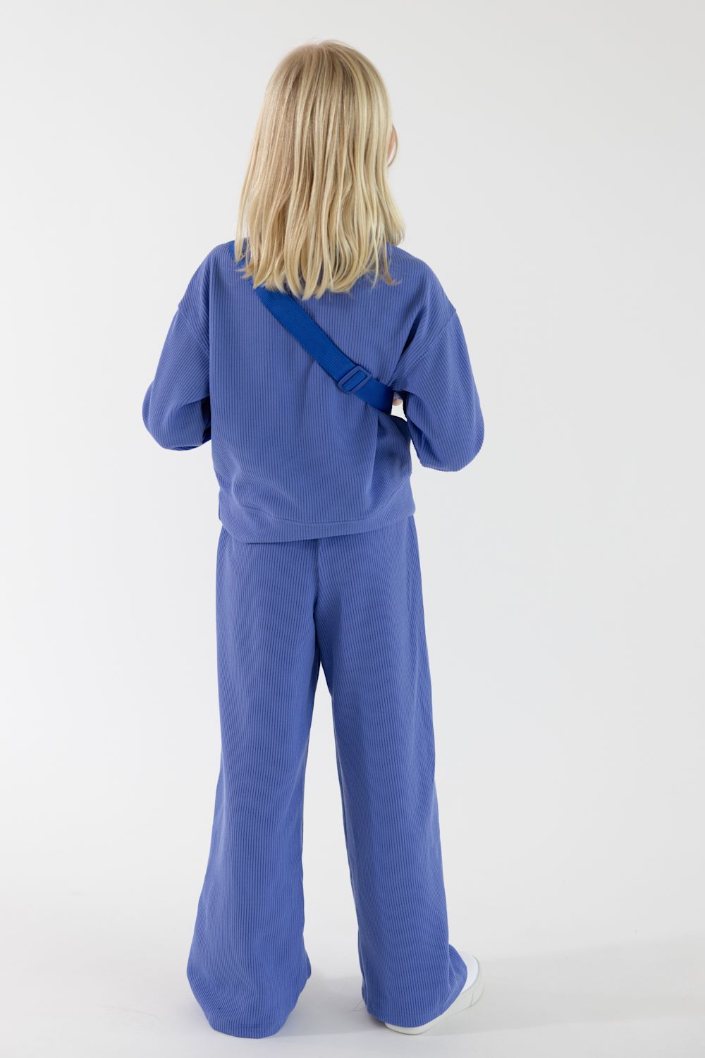 Back view of a young girl with long blonde hair facing away from the camera, wearing a bluish-purple ribbed quarter-zip pullover, matching wide-leg pants, and styled with a matching Perla Blue belt bag slung across her back against a solid white background.
