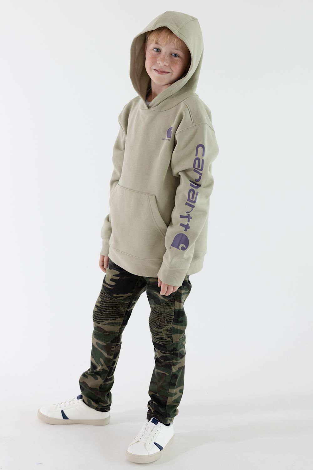 A child wearing a light beige Carhartt hoodie with a purple sleeve logo, paired with camouflage pants and white sneakers, standing against a plain white studio background.