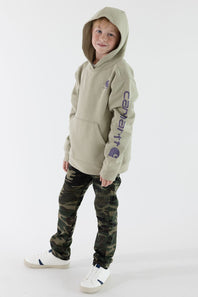 A child wearing a light beige Carhartt hoodie with a purple sleeve logo, paired with camouflage pants and white sneakers, standing against a plain white studio background.