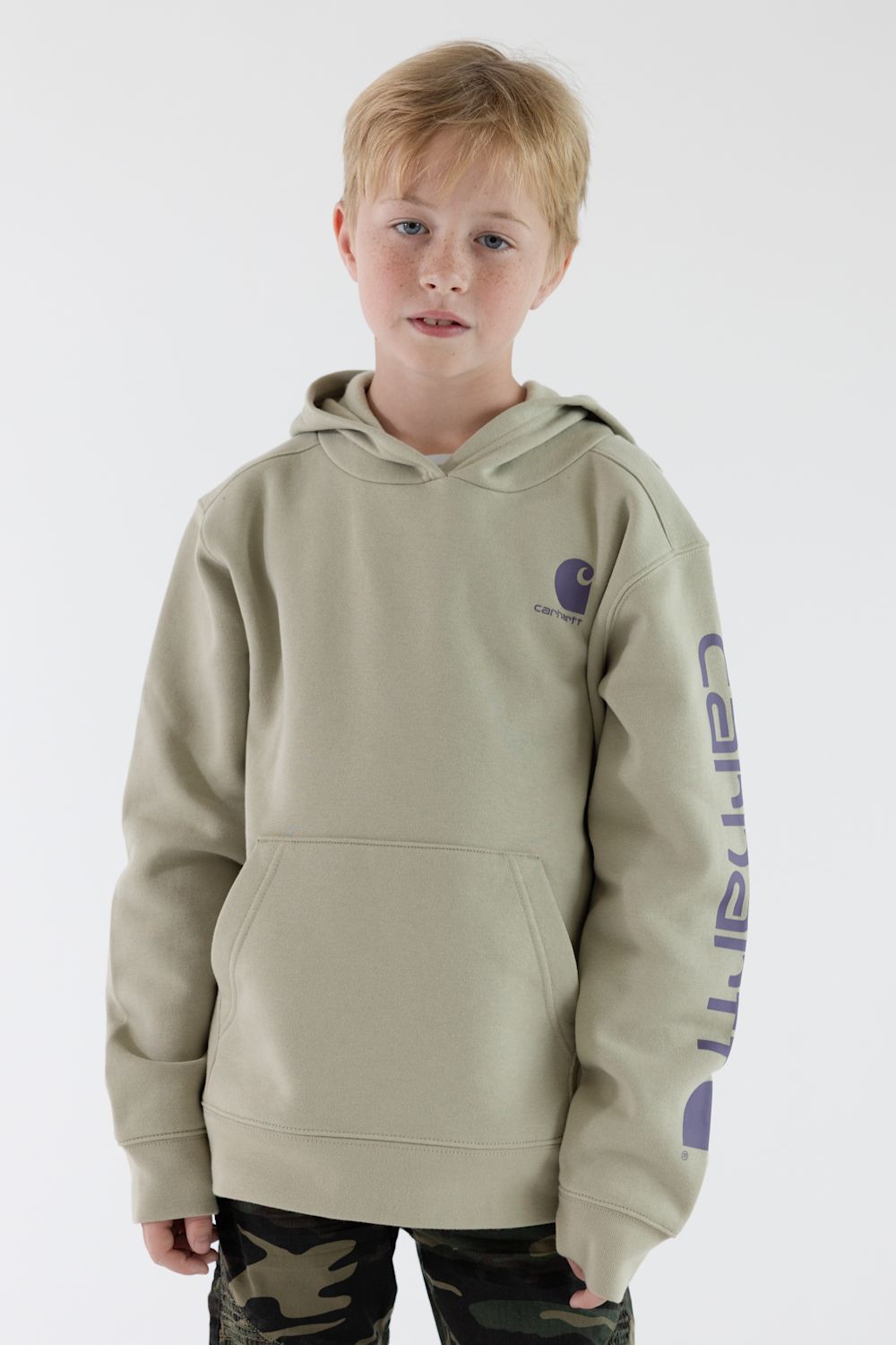 A young boy wearing a light beige Carhartt hoodie with purple branding on the sleeve, paired with camouflage pants, against a plain white studio background.