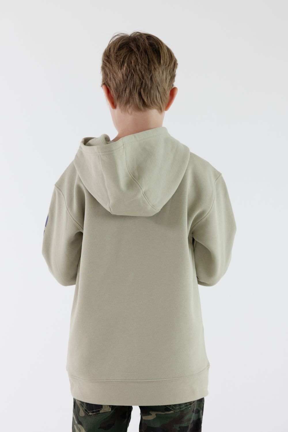 A child viewed from behind wearing a light sage green hooded sweatshirt and camouflage pants against a plain white studio background.