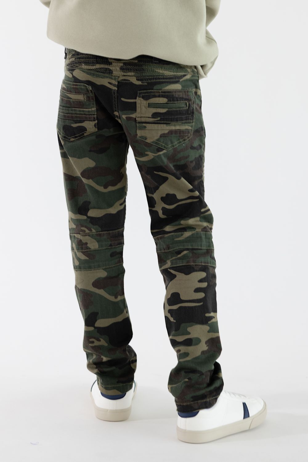 A boy viewed from the back wearing camo denim jeans with a tan shirt and white sneakers in front of a white studio background