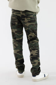 A boy viewed from the back wearing camo denim jeans with a tan shirt and white sneakers in front of a white studio background