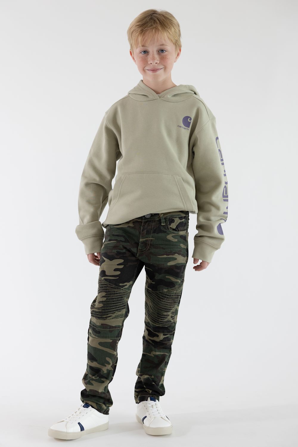 A boy standing in front of a white studio background wearing a tan Carhartt logo hoodie, camo jeans and white sneakers