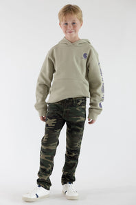 A boy standing in front of a white studio background wearing a tan Carhartt logo hoodie, camo jeans and white sneakers