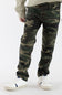 A boy wearing camo print jeans with a tan long sleeve and white shoes standing in front of a clean studio background