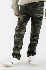 A boy wearing camo print jeans with a tan long sleeve and white shoes standing in front of a clean studio background