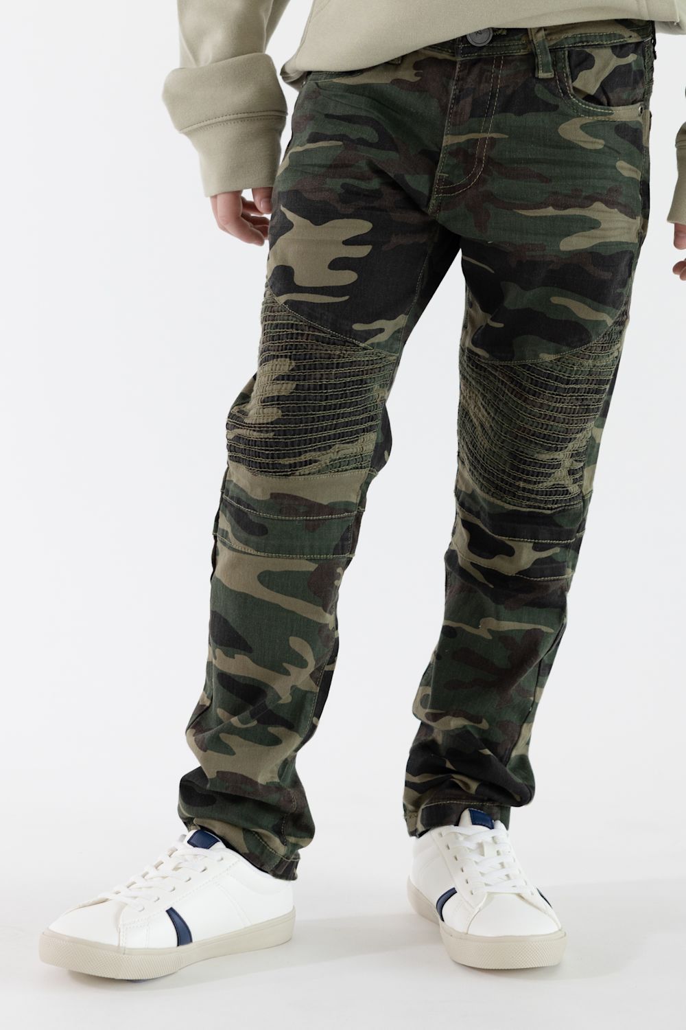 A boy wearing camo print jeans with a tan long sleeve and white shoes standing in front of a clean studio background