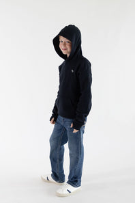 A child wearing a black pullover hoodie, blue jeans, and white sneakers, standing against a plain white studio background.