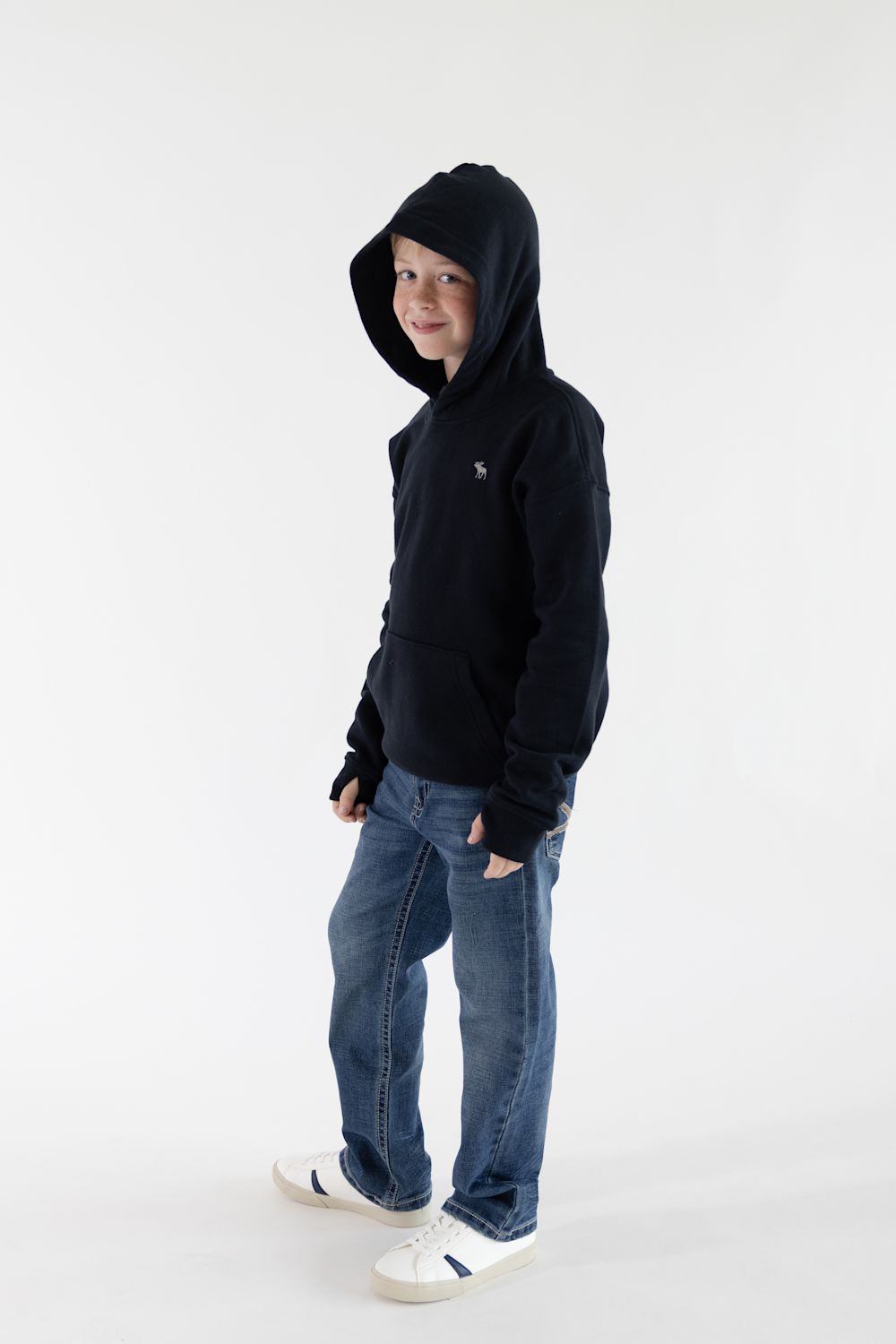 A child wearing a black pullover hoodie, blue jeans, and white sneakers, standing against a plain white studio background.