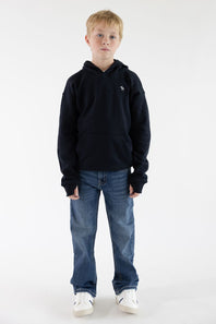 A young boy standing against a white studio background, wearing a dark navy pullover hoodie with a small embroidered logo on the left chest, blue jeans, and white sneakers.