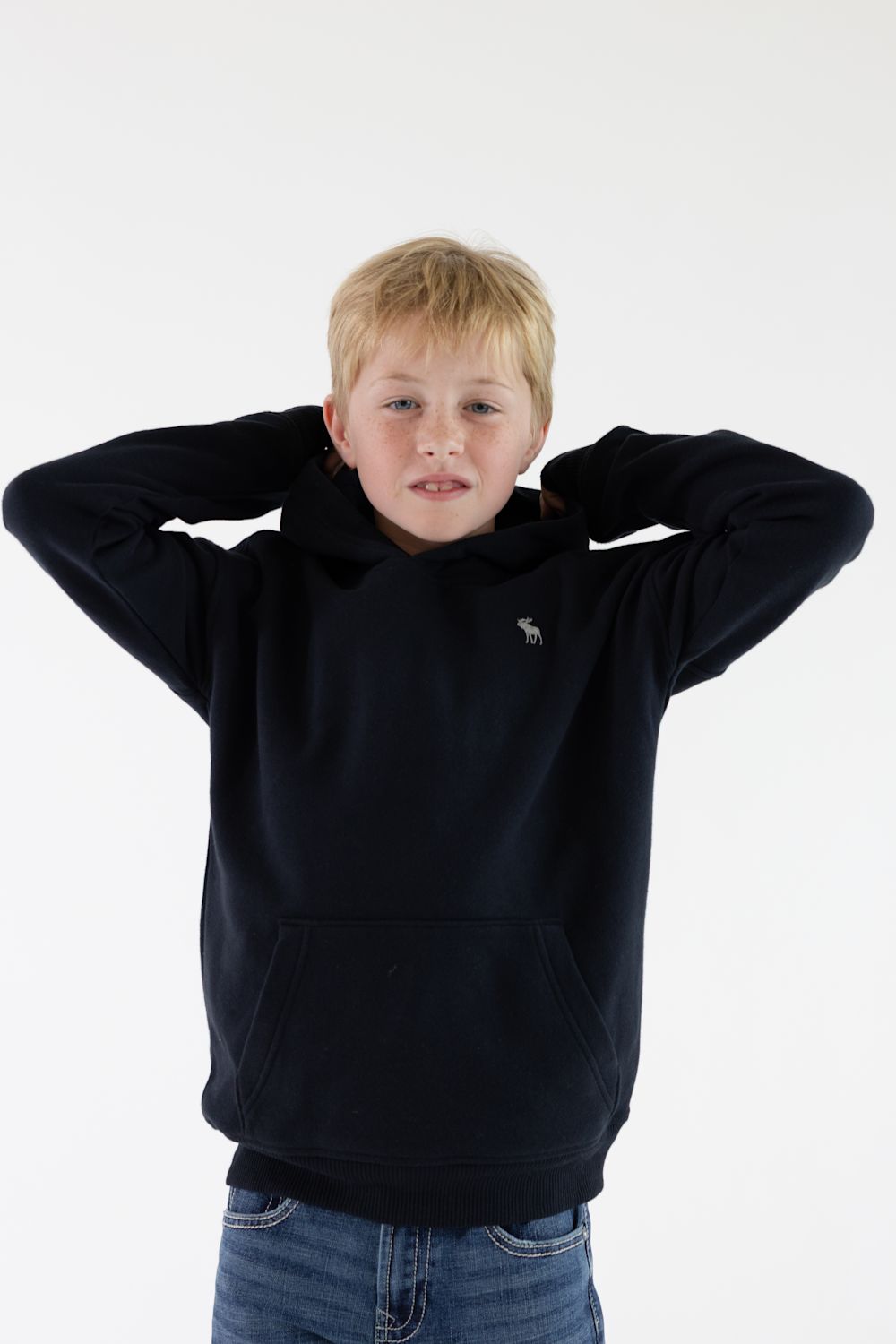 A young boy with blonde hair wearing a dark navy blue hoodie with a small embroidered moose logo on the left chest, paired with blue jeans, posing with his hands behind his head against a plain white background.