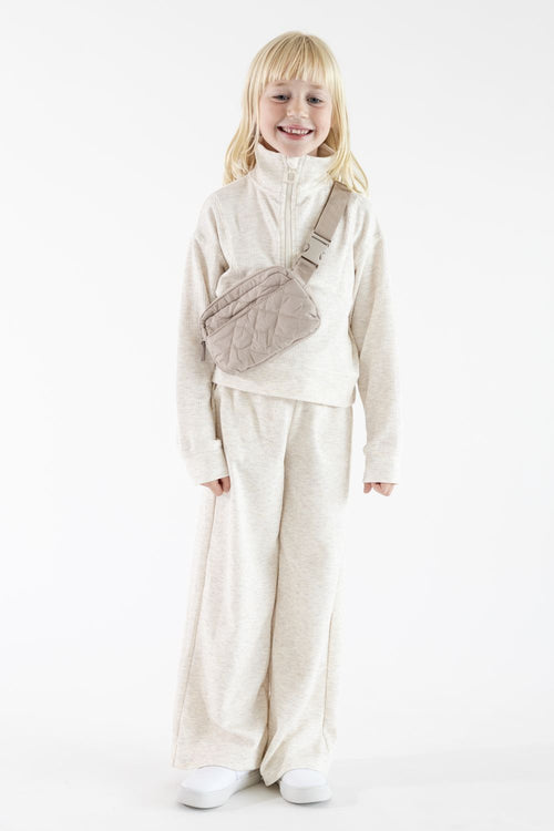A young girl with long blonde hair smiling and standing facing forward, wearing an oatmeal-colored long-sleeve ribbed quarter-zip pullover with matching wide-leg pants, styled with a plush oatmeal belt bag across her torso on a solid white background.