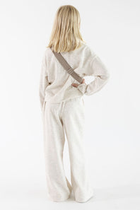 Back view of a young girl with long blonde hair facing away from the camera, wearing an oatmeal-colored long-sleeve ribbed quarter-zip pullover, matching wide-leg pants, and a matching plush belt bag slung across her back against a solid white background.