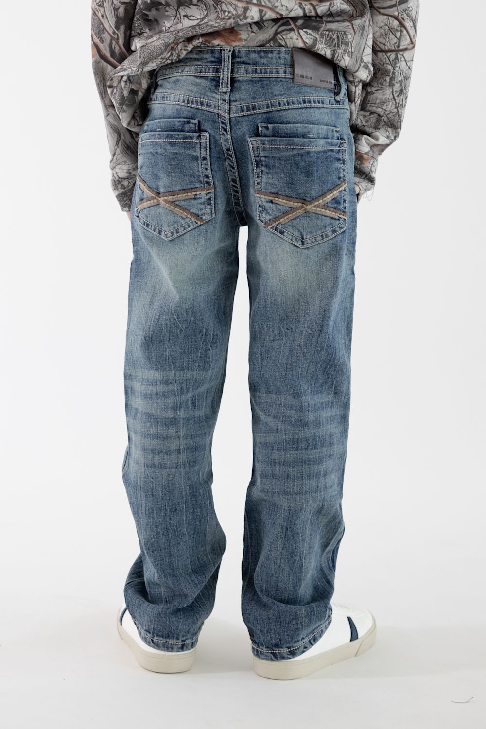 A boy viewed from the back wearing dark wash denim jeans with tan back pocket stitching, a long sleeve camo shirt, and white sneakers on a white studio background