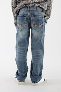 A boy viewed from the back wearing dark wash denim jeans with tan back pocket stitching, a long sleeve camo shirt, and white sneakers on a white studio background