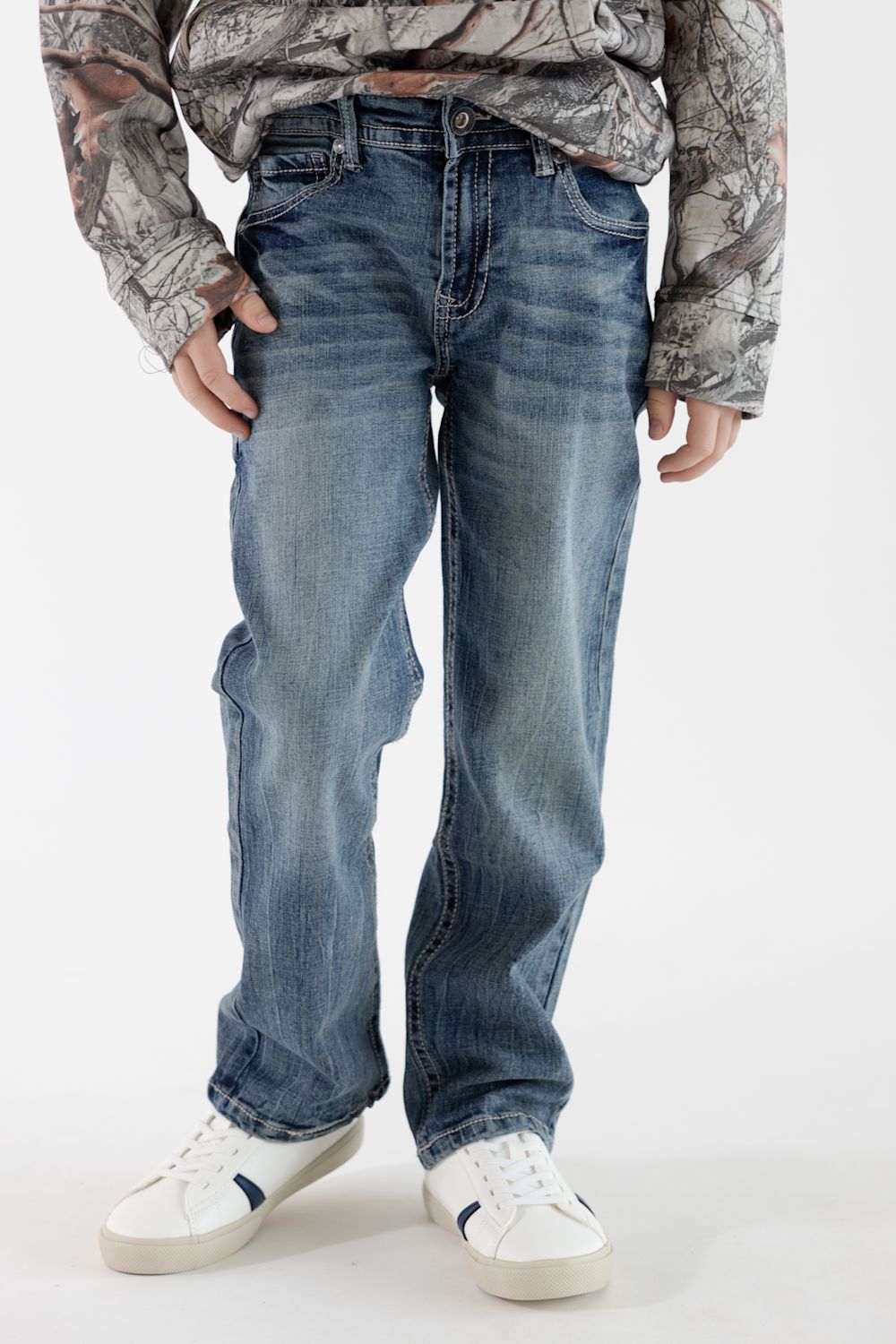 Boy wearing dark wash denim jeans with a long sleeve camo hoodie and white sneakers standing in front of a white studio background