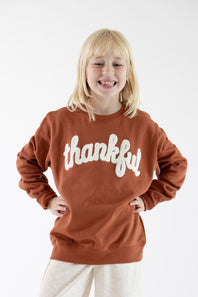 A young girl with blonde hair smiling while wearing a rust-brown crewneck sweatshirt with 'thankful' written across the chest in white script, paired with light beige pants, against a plain white background.