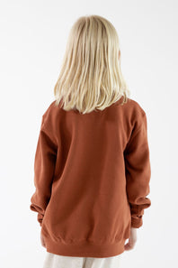 A child with blonde hair seen from the back, wearing a rust-colored crewneck sweatshirt and light-colored pants against a plain white background.