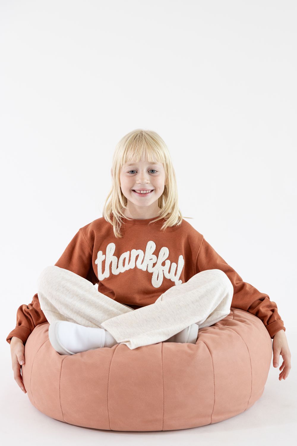 A young girl with blonde hair sits cross-legged on a large dusty pink bean bag chair, wearing a rust-orange sweatshirt with the word 'thankful' in white script and cream-colored pants, against a plain white studio background.