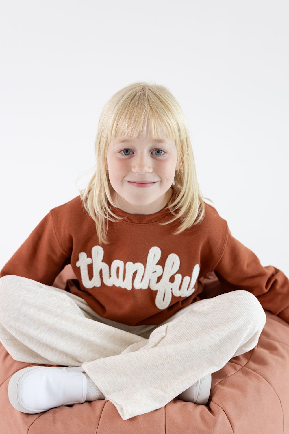 A young blonde girl sitting cross-legged on a dusty rose beanbag chair, wearing a rust-brown sweatshirt with the word 'thankful' in white script across the chest, paired with cream-colored sweatpants.
